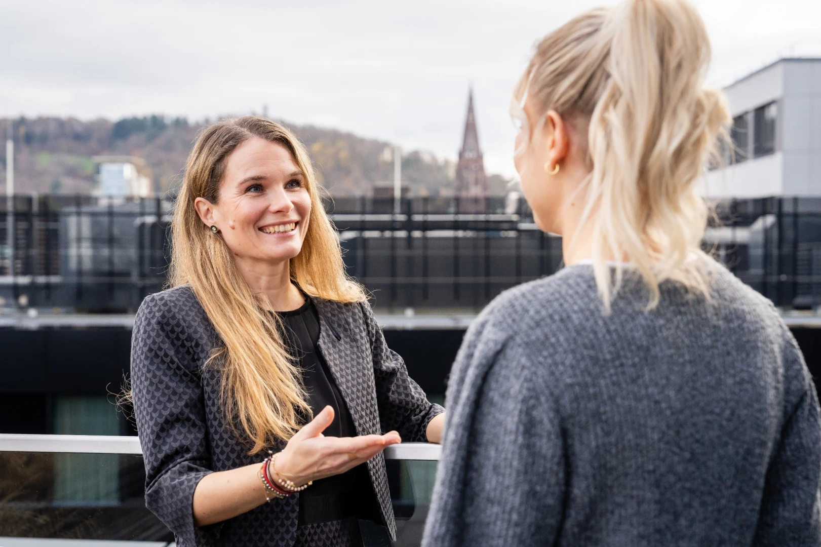 Zwei Personen stehen sich auf einer Dachterrasse vor einer städtischen Kulisse gegenüber und sprechen miteinander, während eine Person mit einer Hand eine erklärende Geste macht.