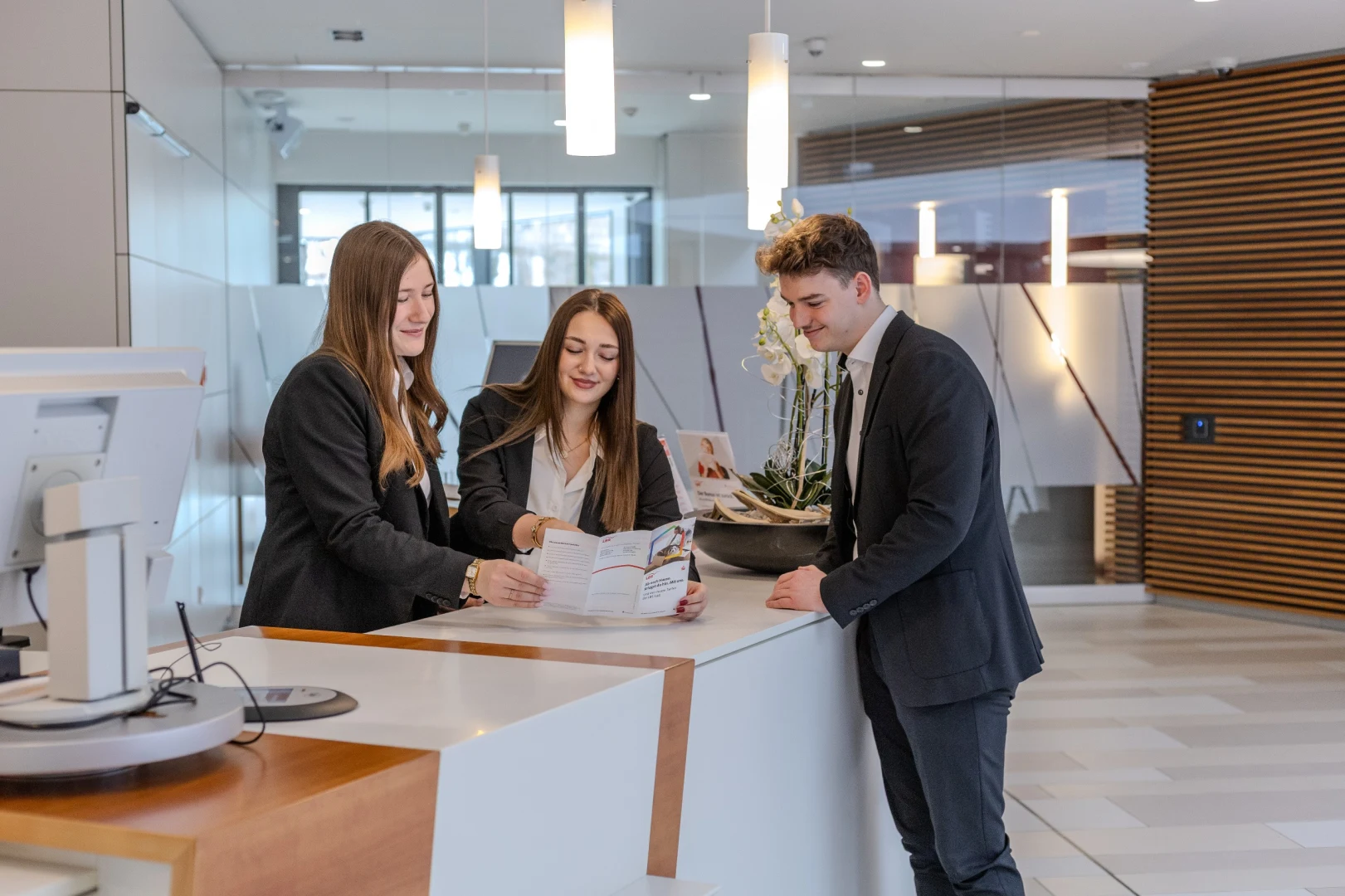 Drei Personen in Business-Kleidung stehen an einem Empfangstresen und betrachten gemeinsam ein Dokument. Der Empfangsbereich ist modern eingerichtet und hell beleuchtet.
